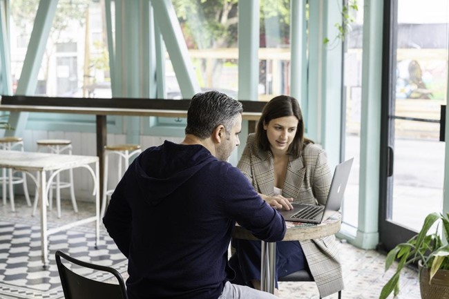 Two people sitting at a table in a cafe, looking at a laptop, discussing account planning.